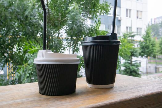 Two black paper coffee cups with lids outdoors in summer in sunny weather on a wooden table of a cafe, coffee shop or restaurant