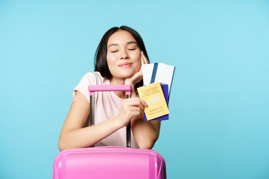 Smiling dreamy asian tourist, girl thinks of travelling, holding passport and tickets, coronavirus international vaccination certificate, going on trip abroad, blue background.