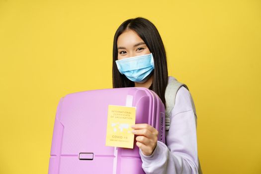 Covid-19 and tourism concept. Happy asian female tourist holding suitcase, showing international coronavirus vaccination certificate for travelling, yellow background.