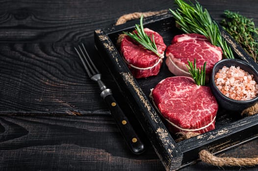 Fillet Mignon tenderloin raw meat veak steaks in wooden tray with herbs. Black wooden background. Top view. Copy space.
