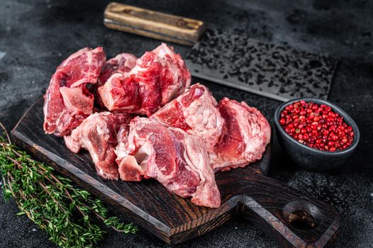 Raw lamb meat stew cuts with bone on wooden butcher board and cleaver. Black background. Top view.
