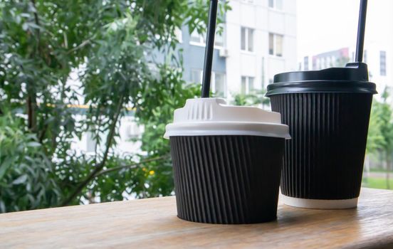 Two black paper coffee cups with lids outdoors in summer in sunny weather on a wooden table of a cafe, coffee shop or restaurant