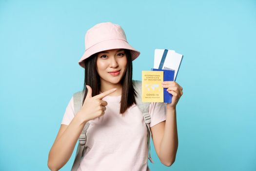 Smiling korean tourist shows her passport, vaccination certificate from coronavirus disease, getting vaccine shot to travel, standing over blue background.