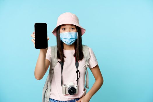 Enthusiastic young asian woman in medical face mask, showing mobile phone app interface, tourism application for travellers, standing voer blue background.