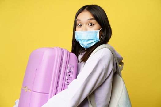 Surprised asian girl tourist, wear medical face mask, looking amazed and impressed at camera, carry suitcase in hands, yellow background.