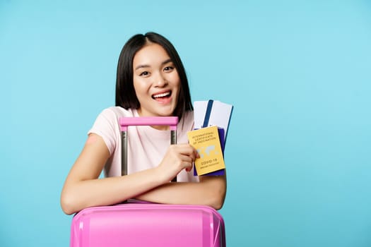 Enthusiastic asian girl shows passport tickets and health covid-19 vaccination certificate, smiling pleased, standing with suitcare, blue background.