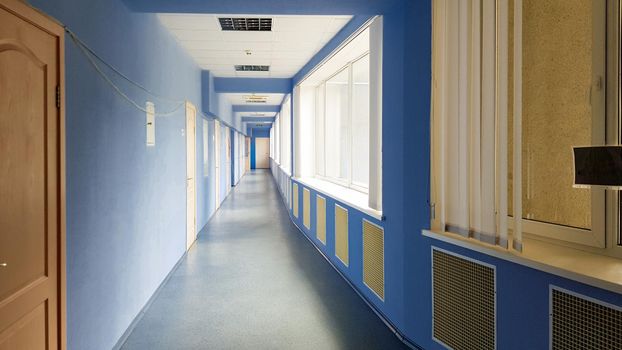 Ukraine, Kiev - September 24, 2019: long empty corridor without people in a hospital with blue walls, wooden doors and windows. Sun rays from the windows in a long corridor of blue