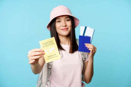 Happy smiling asian tourist, girl shows international vaccination passport, health passport and flight tickets, getting abroad after covid-19 vaccine shot, blue background.