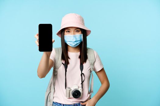 Smiling asian woman, tourist in medical face mask, with camera, showing smartphone empty screen, travelling, recommending tourism app on phone, blue background.