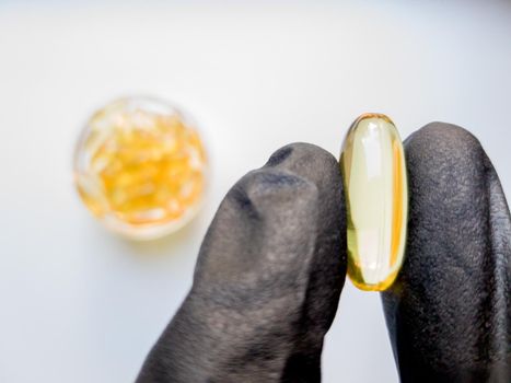 fish oil capsules in hand in a black medical glove on a white background, the hand takes one tablet from a saucer or plate. Hand holding omega 3 capsule on a white background. Healthcare concept.
