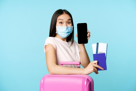 Enthusiastic asian girl, vaccinated tourist standing with cute suitcase, flight tickets and passport, showing vaccination certificate, health info on smartphone screen, wearing face mask.