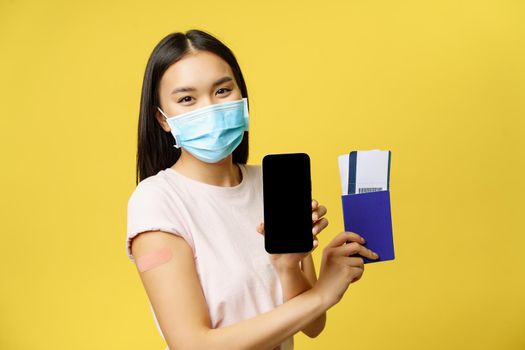 Covid-19 and tourism concept. Smiling asian woman vaccinated, showing smartphone screen, app interface, passport with tickets for vacation, yellow background.