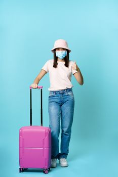 Vertical shot of vaccinated asian girl in medical face mask, going on vacation, standing with suitcase and showing thumb up, blue background.