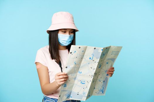 Asian girl tourist in medical face mask, vaccinated from covid with patch on arm, travelling abroad, looking at sightseeing map, exploring tourism spots, standing over blue background.