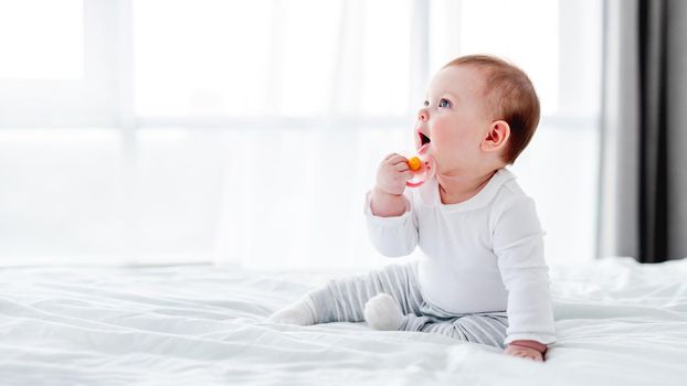 Adorable little baby boy sitting in the bed and holding dummy in his hand close to his mouth. Beautiful toddler child in the bedroom in the morning time. Cute kid alone at home