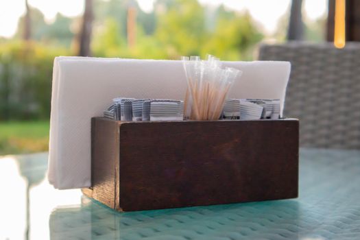 A brown wooden napkin holder and toothpicks sit on the glass table of the cafe, illuminated by the sun's rays. Table setting in the restaurant. Glass table in a green outdoor garden