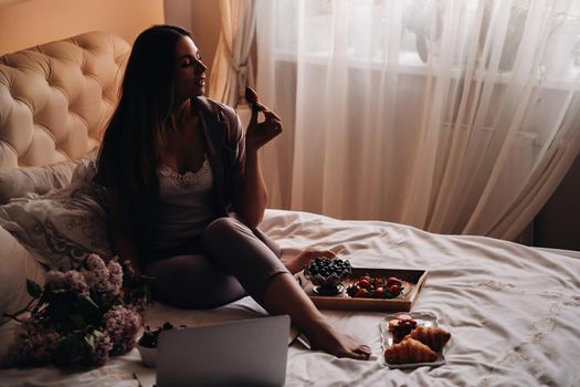 a girl sits in bed in the evening watching a laptop and eating strawberries, a girl in bed eats sweets before going to bed.