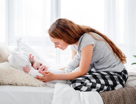 Beautiful young mother sitting in the bed and looking at her adorable little baby boy. Pretty smiling girl with kid child in morning time. Happy matherinity moments