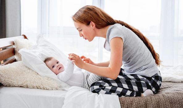 Beautiful young mother holding hands of her adorable little baby boy and looking at the child. Pretty girl with kid in the bed in morning time. Happy family moments