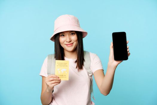 Smiling asian girl shows international covid-19 vaccination certificate, health passport on smartphone screen, tourism app for travellers, standing over blue background.