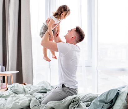 Young father sitting in the bed and spending morning with his little daughter holding her in his hands against the window. Smiling child with her parent in sunny room with stylish interior