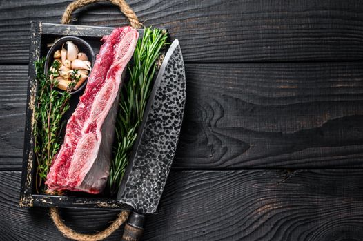 Fresh Raw veal short ribs in a wooden tray with herbs. Black wooden background. Top view. Copy space.