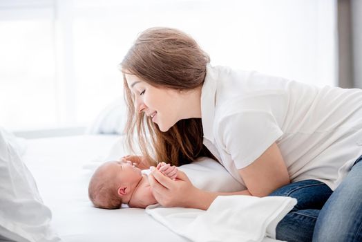 Pretty young mother holding hands of her sleeping newborn baby daughter in the bed and looking at her with smile. Parent and infant child. Happy moments of matherinity