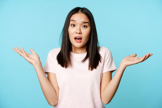 Confused asian girl spread empty hands sideways, shrugging shoulders clueless, standing in tshirt over blue background.