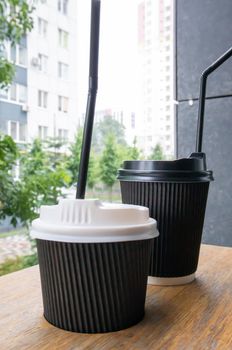 Two black paper coffee cups with lids outdoors in summer in sunny weather on a wooden table of a cafe, coffee shop or restaurant
