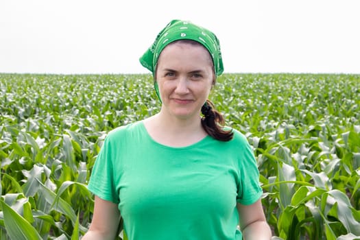 Beautiful smiling young woman farmer in green t-shirt and green sun hat standing in the middle of a green corn field on a sunny summer day outdoors. Harvest time. Organic farming. Sunlight