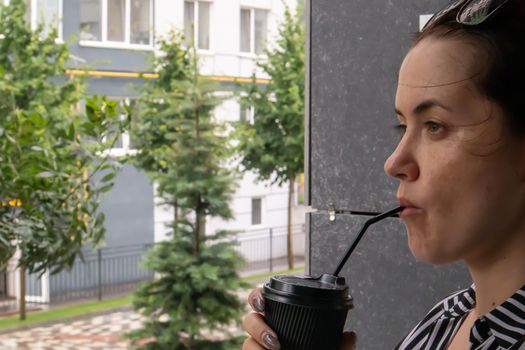 One black paper cup with a lid and a straw in the hands of a young happy smiling woman outdoors in summer in sunny weather on the background of houses, in a coffee shop or restaurant. Coffee to go