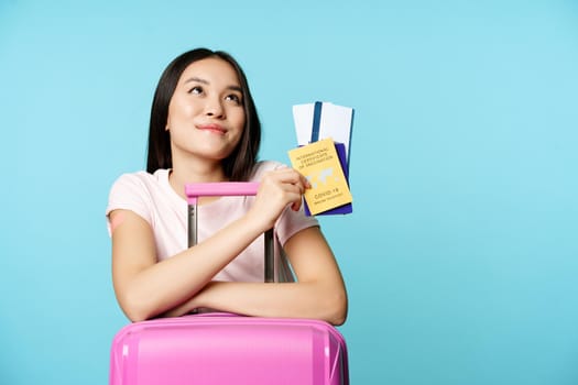 Happy asian female tourist dreaming of trip, lean on suitcase, holding covid-19 international vaccination certificate, two tickets and passport, thinking of journey, blue background.