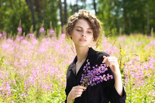 A young woman with a bouquet of lupines in her hand against the background of blooming Ivan-tea on a bright sunny summer day