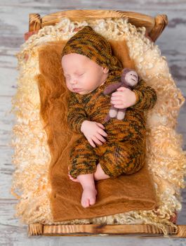 newborn sleeping boy on little bed in brown knitted suit and hat with toy in hand