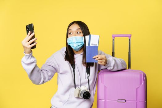 Excited asian tourist, girl travelling, taking selfie on smartphone with flight tickets and passport, going on vacation and posting photo on social media, yellow background.