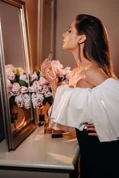 A beautiful girl paints her lips in the reflection of the mirror at home. A girl looks in the mirror before the holiday and holds a lipstick in her hands. Girl with long hair goes to a party.