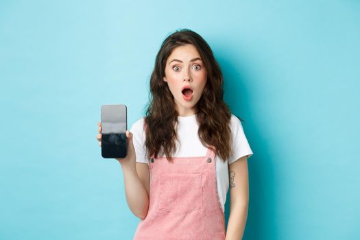 Shocked beautiful girl drop jaw and show empty smartphone screen, demonstrate your app or online store on blank phone display, standing against blue background.