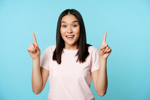 Enthusiastic korean girl shows advertisement, points up and smiles amazed, checks out promo deal, stands over blue background.