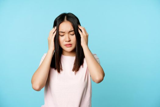 Asian young girl with headache, massaging her head with troubled face expression, painful migraine, standing over blue background.