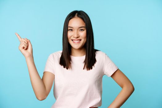 Cute smiling asian brunette, student pointing at upper left corner, showing company name, promo banner, blue background.