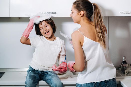 housewife mom in pink gloves washes dishes with her son by hand in the sink with detergent. A girl in white and a child with a cast cleans the house and washes dishes in homemade pink gloves.A child with a cast washes dishes and smiles.