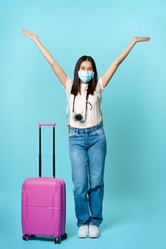Full length shot of happy asian girl tourist, wearing face medical mask, standing near cute suitcase, going on vacation during covid pandemic, excited about travelling abroad.