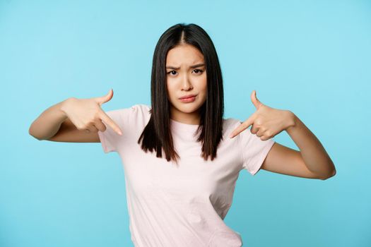 Sassy and cool asian woman pointing fingers at logo, showing smth and furrow eyebrows complicated, standing over blue background.