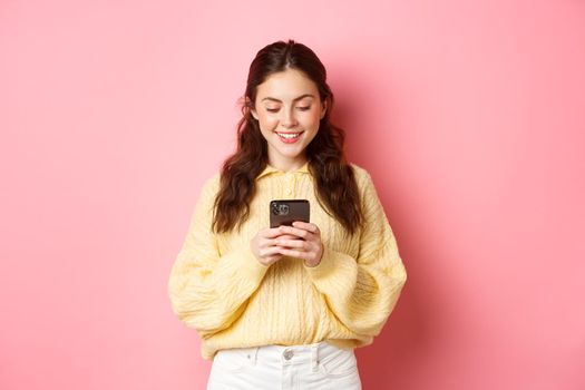 Portrait of young brunette woman reading smartphone screen and smiling, chatting on social media app, shopping online, standing against pink background.