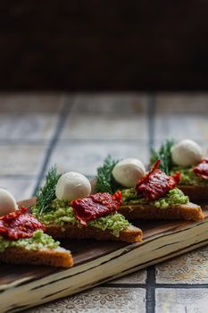 Mini triangle canapes bruschetta with avocado, mozarella and dried tomatos on the rustic board on the vintage tiled background.