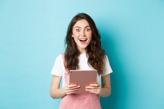 Surprised happy woman stare at camera amazed, hear amazing news, holding tablet in hands, standing over blue background.