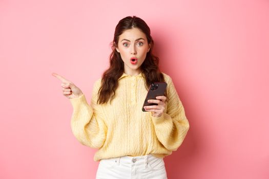 Surprised young beautiful girl gasping and saying wow, pointing finger aside at left copyspace, holding mobile phone, standing against pink background.