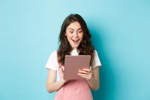 Image of excited young woman stare surprised at digital tablet screen, found something cool online, standing against blue background.