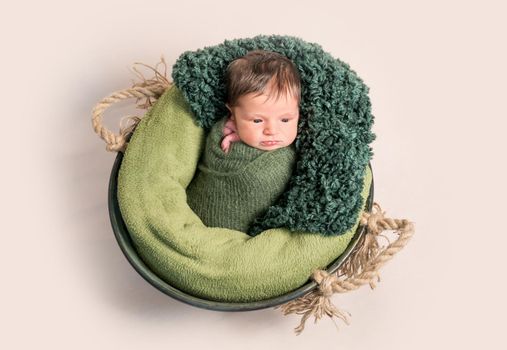 Little newborn baby swaddled in green coccon lying on round basket