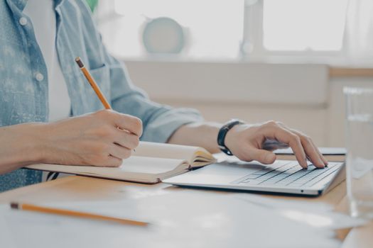 Close up photo of male hands typing on computer keyboard and writing notes in agenda, freelance worker typing message and contacting client by internet from home while sitting at kitchen table